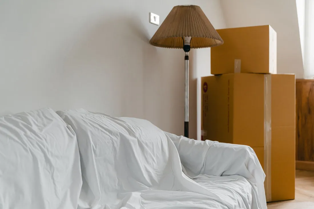 Weiß abgedecktes Sofa neben Stehlampe und gestapelten Umzugskartons in einem hellen Zimmer