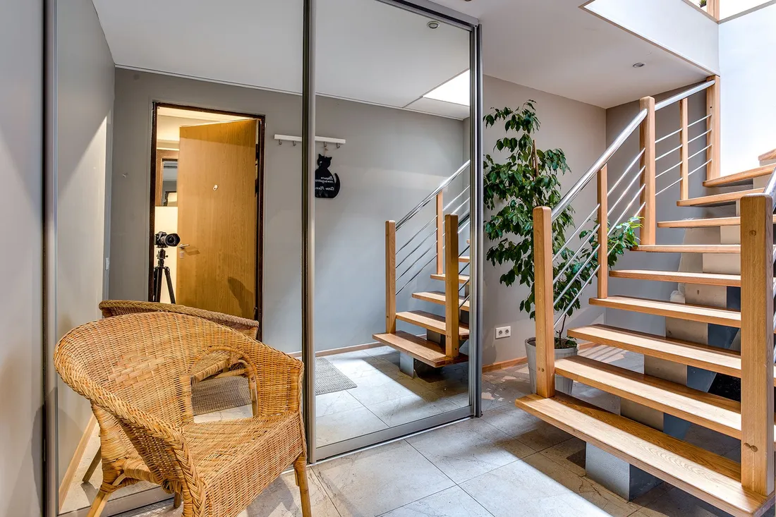 Hausflur mit Korbsessel, verspiegelter Schranktür und offener Holztreppe neben einer großen Zimmerpflanze