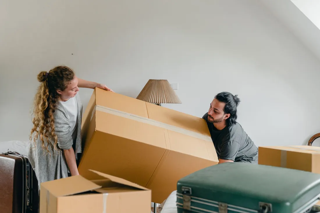 Frau und Mann heben gemeinsam einen großen Umzugskarton in einem hellen Dachzimmer an