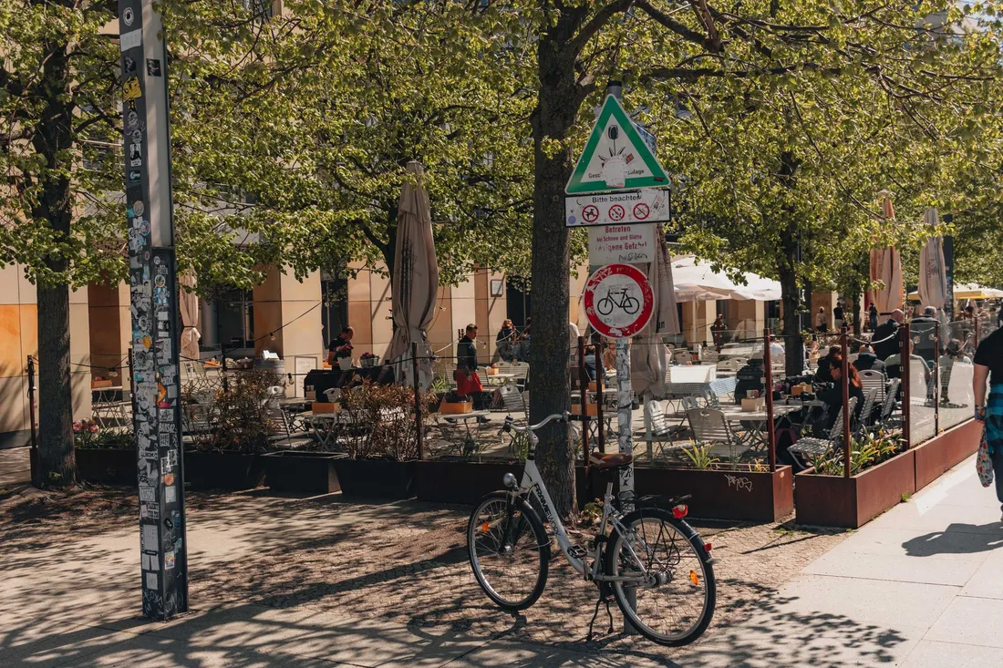 Fahrrad an einem beklebten Schildermast vor einer Caféterrasse unter hohen Bäumen