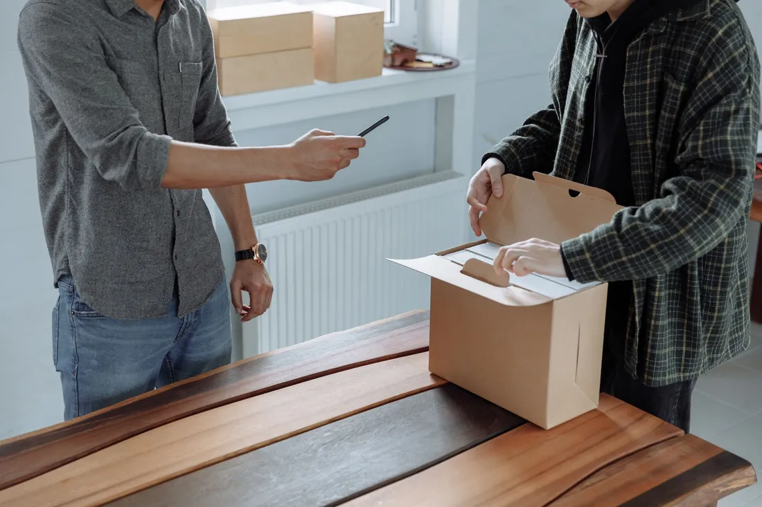 Zwei Männer bauen an einem Holztisch einen Umzugskarton zusammen, einer hält einen Stift